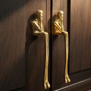 May include: Two gold-colored cabinet handles in the shape of a person sitting with their chin resting on their hand. The handles are mounted on a dark brown wooden cabinet door. The handles have a shiny, reflective surface.