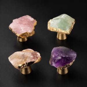 May include: Four decorative cabinet knobs with natural stone faces in pink, green, tan, and purple. Each knob has a gold-colored trim and a brass-colored base. The knobs are arranged on a black surface.