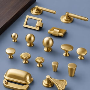 May include: A collection of various gold-colored cabinet hardware, including knobs, pulls, and handles. The hardware features different shapes and designs, such as round, square, and fluted styles. The items are arranged on a light blue surface.