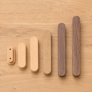 May include: Six wooden cabinet pulls arranged in a row by size. The first four pulls are light-colored wood with rounded ends, and the last two are dark-colored wood with rounded ends. The smallest pull shows two screw holes on its front surface.