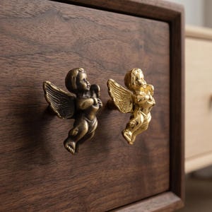 May include: Two decorative drawer pulls in the shape of cherubs. One pull is bronze-colored, and the other is gold-colored. The pulls are attached to a dark brown wooden drawer. The cherubs have detailed wings and are holding an object.