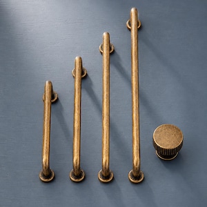May include: A collection of antique brass cabinet hardware, including four pull handles of varying lengths and a round knob. The hardware has a distressed finish and is arranged on a blue-gray surface.
