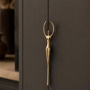 May include: A gold-colored cabinet pull in the shape of a stylized human figure with arms raised. The pull is mounted on a dark gray cabinet door. The design is elegant and modern, suitable for various interior styles.