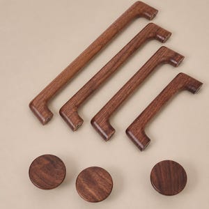 May include: A collection of four dark brown wooden cabinet pulls of varying lengths arranged diagonally, accompanied by three circular wooden cabinet knobs. Each piece features a visible wood grain pattern and a smooth, polished finish.