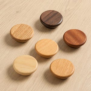 May include: Six wooden cabinet knobs in various shades of brown and tan are arranged on a light wood surface. The knobs are circular with a slightly rounded shape, showcasing the natural wood grain.