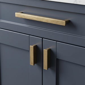 May include: Close-up of a navy blue cabinet with gold-toned hardware. The cabinet features a long, rectangular handle on the top drawer and two smaller, rectangular handles on the cabinet doors below. A white marble countertop is visible.