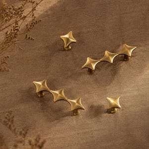 May include: A collection of gold-toned metal hardware including two single-point knobs and two multi-point drawer pulls. Each piece features a four-pointed star shape with a raised center, finished in a smooth, matte metallic texture.