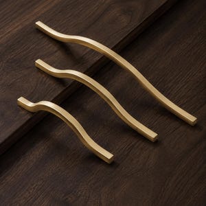 May include: Three brass-colored cabinet handles with a wavy design are arranged diagonally on a dark wood surface. The handles vary in size, with the largest at the top and the smallest at the bottom. The handles have a smooth, matte finish.