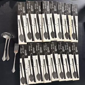 May include: A collection of sixteen black rectangular boxes labeled "Fine Stainless Tableware by Oneida Ltd." arranged in two rows, alongside two silver-toned spoons and one silver-toned fork. Each box features a printed illustration of a utensil.