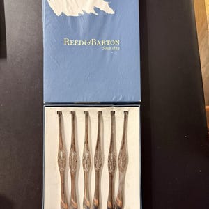 May include: A set of six silver-toned metal seafood picks with lobster motifs on the handles, presented in a blue Reed & Barton box. The box lid features the text Reed & Barton Since 1824 in gold lettering against a blue background with a white graphic.