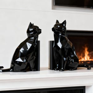 May include: Two black geometric cat sculptures sit side-by-side on a white surface. The cat figures have a faceted design, giving them a modern look. The background includes a fireplace with a burning fire.