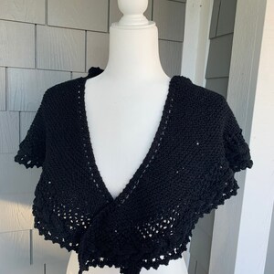 May include: A black knitted shawl with a delicate lace trim. The shawl is draped over a white mannequin.
