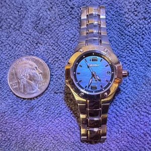 Puede incluir: Un reloj de pulsera Seiko dorado y plateado con esfera azul iridiscente. El reloj tiene una pantalla de fecha y una correa de metal. Una moneda de veinticinco centavos estadounidense está al lado del reloj para comparar el tamaño.