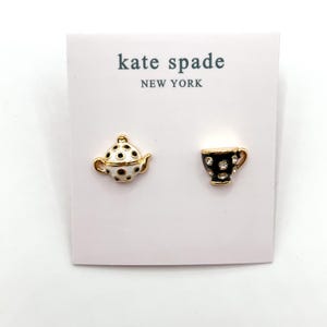 Op de afbeelding: Kate Spade oorbellen met een theepot- en theekopontwerp. De theepot is wit met zwarte stippen en gouden randen. De theekop is zwart met kristallen accenten en gouden randen. Op een roze kaart met het Kate Spade New York logo.