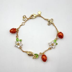 Peut inclure: Bracelet doré orné de breloques fraises rouges, de fleurs blanches et d'un charm en forme d'escargot. Le bracelet est doté d'un fermoir mousqueton et est orné d'accents en cristal transparent. Un bijou charmant.