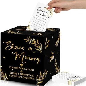 May include: A black square memory box featuring gold foil olive branch illustrations and the text Share a Memory. Below this, the text reads Please take a card and share a memory or a word of love here. A white card with matching text is being inserted.