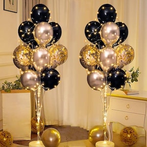 May include: Two balloon bouquets with black, gold, and silver balloons, illuminated by string lights. The balloons are arranged on stands with round bases, and additional gold balloons are on the floor. The overall theme is festive and celebratory.