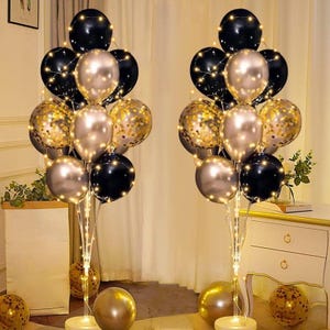 May include: Two balloon bouquets with black, gold, and silver balloons, illuminated by string lights. The balloons are arranged on stands with round bases, and additional gold balloons are on the floor. The overall theme is festive and celebratory.