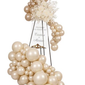 May include: A wedding sign with a floral arrangement and balloon decor. The sign reads "Welcome to Our Wedding Day" and features the names "Cristina & Marian". The balloons are in shades of gold and ivory.