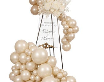 May include: A wedding sign with a floral arrangement and balloon decor. The sign reads "Welcome to Our Wedding Day" and features the names "Cristina & Marian". The balloons are in shades of gold and ivory.