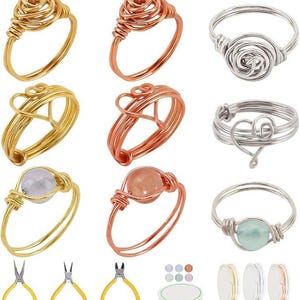 May include: A collection of handmade wire rings in gold, copper, and silver tones. The rings feature various designs, including floral, heart, and gemstone accents. Jewelry-making tools and wire spools are also visible.