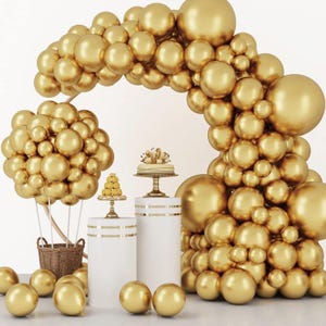 May include: A golden balloon arch and cluster arrangement, with a hot air balloon basket, white pedestals, and a small cake. The scene is decorated with gold balloons and accents, creating a festive and celebratory atmosphere.