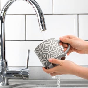 May include: A gray ceramic mug with a white floral pattern is held under a running faucet. The mug has a curved handle and is being washed in a stainless steel sink. The background features white subway tiles.