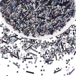May include: A collection of assorted glass beads in shades of silver, black, dark blue, and iridescent purple. The mix includes small round seed beads and elongated tube-shaped bugle beads, some of which are scattered outside a white bowl.
