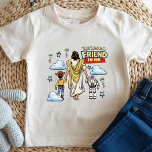 A white t-shirt with a graphic design featuring a cartoon image of Jesus Christ and two children, along with the text "You're a FRIEND in ME" printed on it. The t-shirt is placed on a woven placemat, and there is a crocheted stuffed animal and a pair of blue jeans next to it.
