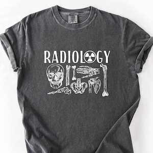 May include: A dark gray t-shirt with the word "RADIOLOGY" in white, along with a graphic of a skull, bones, and a radiation symbol. The shirt is made of a soft material and has rolled-up sleeves. The word "PEPPER" is printed in black at the bottom right.