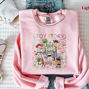 A pink t-shirt with a colorful graphic of characters from the Toy Story movie, including Woody, Buzz Lightyear, and other beloved characters, is displayed on a bed.
