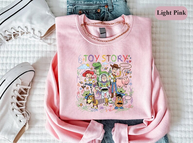 A pink t-shirt with a colorful graphic of characters from the Toy Story movie, including Woody, Buzz Lightyear, and other beloved characters, is displayed on a bed.