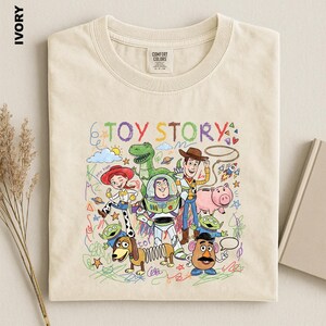 A beige t-shirt with a colorful and whimsical illustration of characters from the Toy Story movie, including Woody, Buzz Lightyear, and other beloved characters.