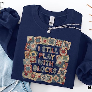 May include: Navy blue sweatshirt with the text "I STILL PLAY WITH BLOCKS" surrounded by a patchwork quilt design. The sweatshirt has long sleeves and a crew neck. A silver necklace with a circular pendant is draped across the sweatshirt.
