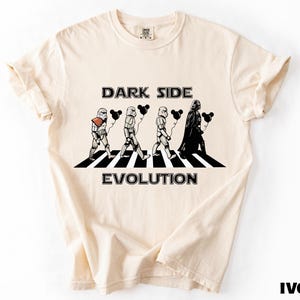 May include: Ivory-colored t-shirt featuring a graphic of Star Wars characters crossing a street, with the text "DARK SIDE EVOLUTION". The design includes Stormtroopers and Darth Vader holding black heart-shaped balloons. The shirt is made of a soft material.
