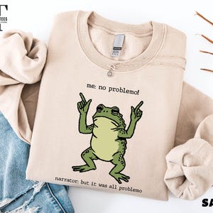 a beige sweatshirt with a cartoon frog graphic and text, along with a denim jacket and some leaves.