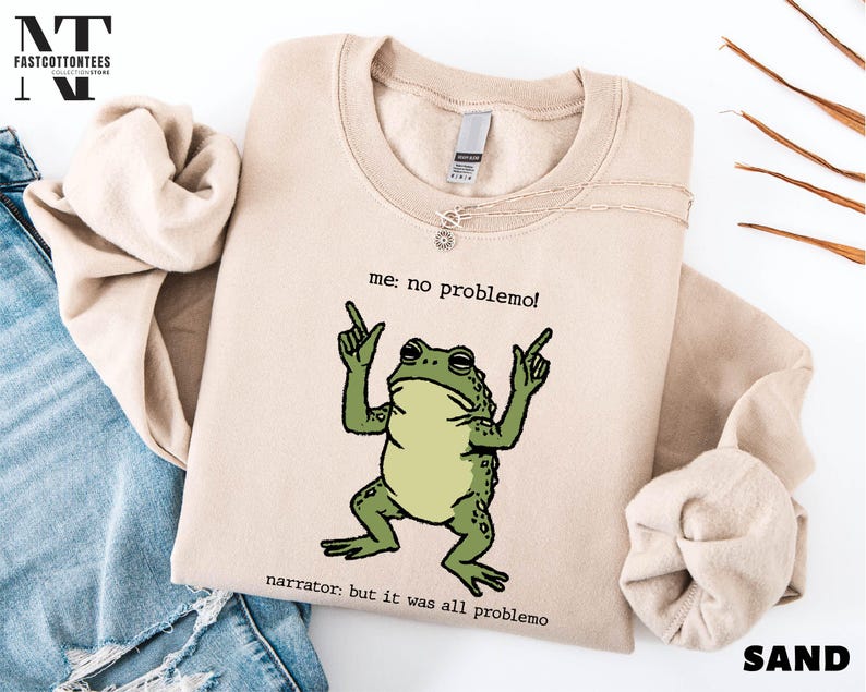 a beige sweatshirt with a cartoon frog graphic and text, along with a denim jacket and some leaves.