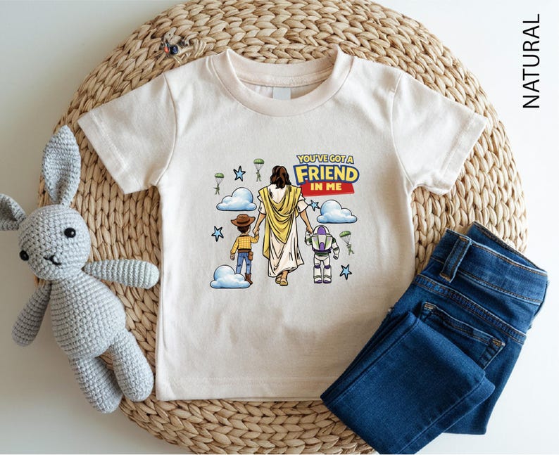A white t-shirt with a graphic design featuring a cartoon image of Jesus Christ and two children, along with the text "You're a FRIEND in ME" printed on it. The t-shirt is placed on a woven placemat, and there is a crocheted stuffed animal and a pair of blue jeans next to it.