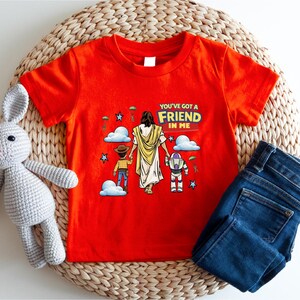 A red t-shirt with a cartoon illustration of a person holding a dog and a robot, along with the text "You've got a FRIEND in me" printed on it. The t-shirt is placed on a woven mat, and there is a stuffed rabbit and a pair of blue jeans next to it.