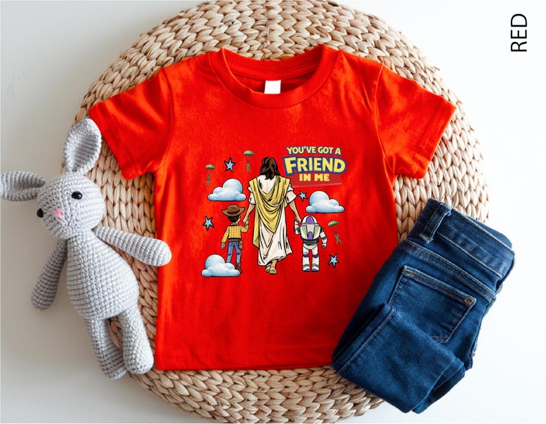 A red t-shirt with a cartoon illustration of a person holding a dog and a robot, along with the text "You've got a FRIEND in me" printed on it. The t-shirt is placed on a woven mat, and there is a stuffed rabbit and a pair of blue jeans next to it.