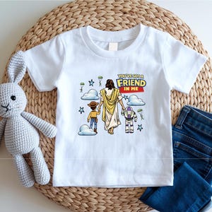 A white t-shirt with a graphic design featuring a cartoon character and the text "You've got a FRIEND in me" is displayed on a woven placemat. Next to the t-shirt are a crocheted rabbit toy and a pair of blue jeans.