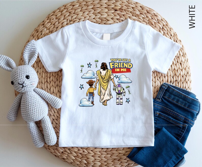 A white t-shirt with a graphic design featuring a cartoon character and the text "You've got a FRIEND in me" is displayed on a woven placemat. Next to the t-shirt are a crocheted rabbit toy and a pair of blue jeans.