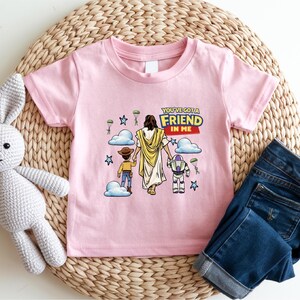 A pink t-shirt with a cartoon illustration of a person holding a child and a robot, along with the text "You've got a friend in me" and a Bible verse. The t-shirt is placed on a woven placemat, and there is a crocheted stuffed rabbit next to it.