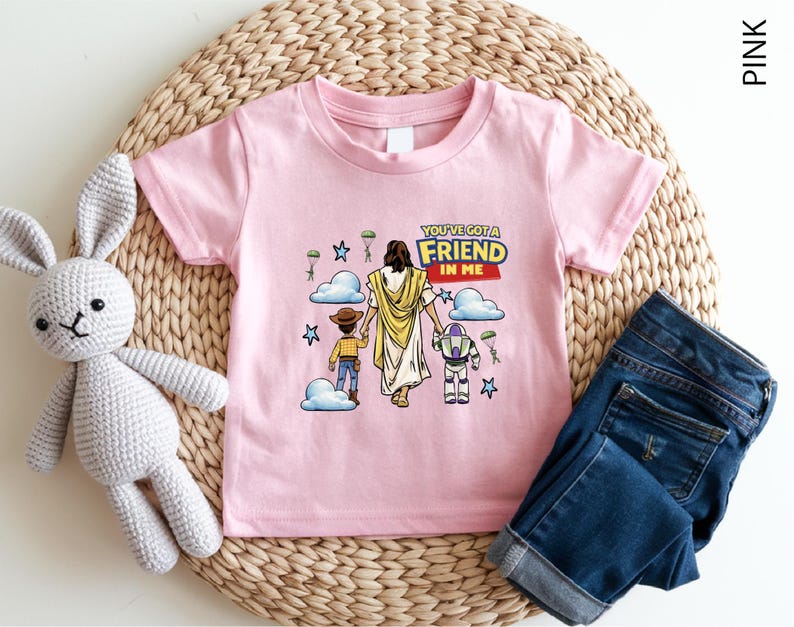 A pink t-shirt with a cartoon illustration of a person holding a child and a robot, along with the text "You've got a friend in me" and a Bible verse. The t-shirt is placed on a woven placemat, and there is a crocheted stuffed rabbit next to it.