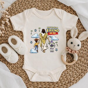A baby onesie with a cartoon image of Jesus Christ and a message, along with a crocheted rabbit toy and a pair of white booties, all arranged on a woven mat.