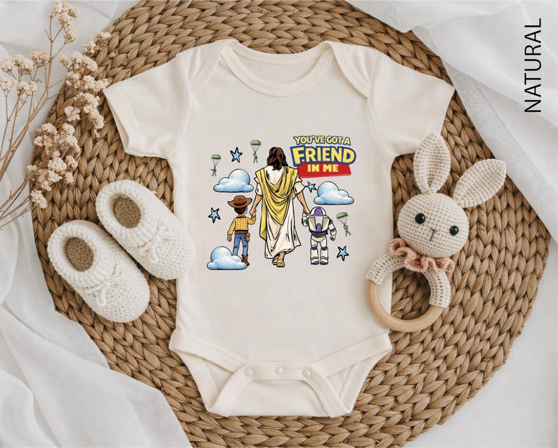 A baby onesie with a cartoon image of Jesus Christ and a message, along with a crocheted rabbit toy and a pair of white booties, all arranged on a woven mat.