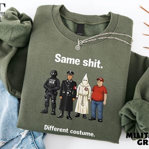 May include: Military green sweatshirt with the text "Same shit. Different costume." and illustrations of four figures. A silver necklace is draped across the sweatshirt. The sweatshirt has long sleeves and a crew neck.