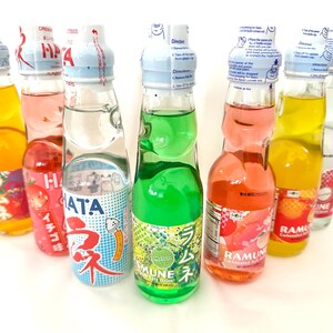 May include: A collection of colorful Ramune soda bottles. The glass bottles contain various fruit-flavored sodas, including orange, green, pink, and clear. Each bottle has a unique label with Japanese text and a marble-style stopper.