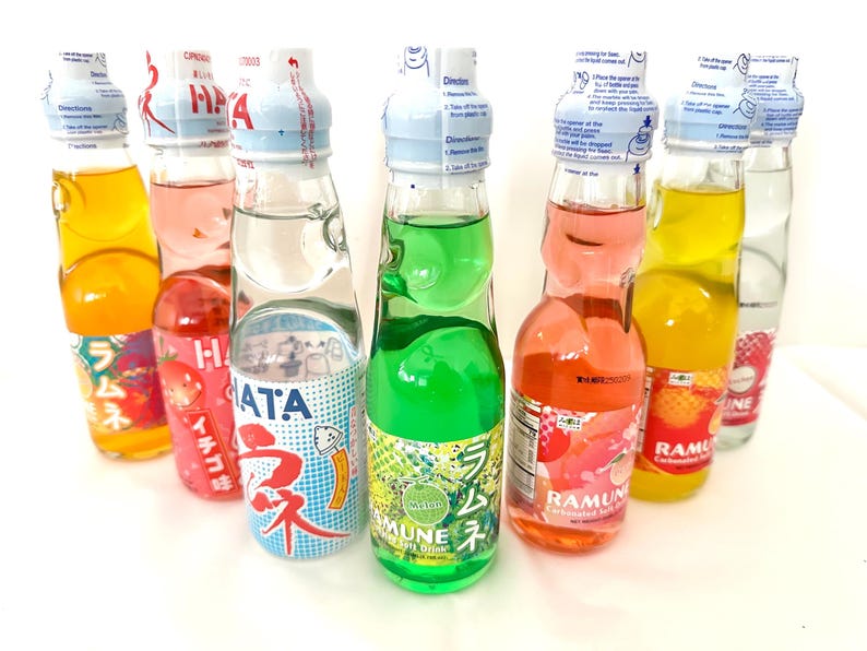 May include: A collection of colorful Ramune soda bottles. The glass bottles contain various fruit-flavored sodas, including orange, green, pink, and clear. Each bottle has a unique label with Japanese text and a marble-style stopper.