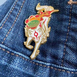 May include: A gold-colored enamel pin featuring a white fox with red markings. The fox is standing on its hind legs with its tail curled up. The fox is wearing a green and red hat with a gold trim. The pin is on a blue denim background.